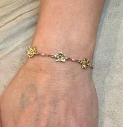 Close-up of wrist wearing a dainty gold floral bracelet with three flower charms and tiny pink beads on a light fabric background