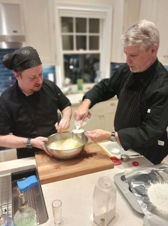 Two cooks in a modern home kitchen whisking batter in a large stainless steel bowl while one pours milk from a measuring cup on a wooden cutting board - home baking prep.