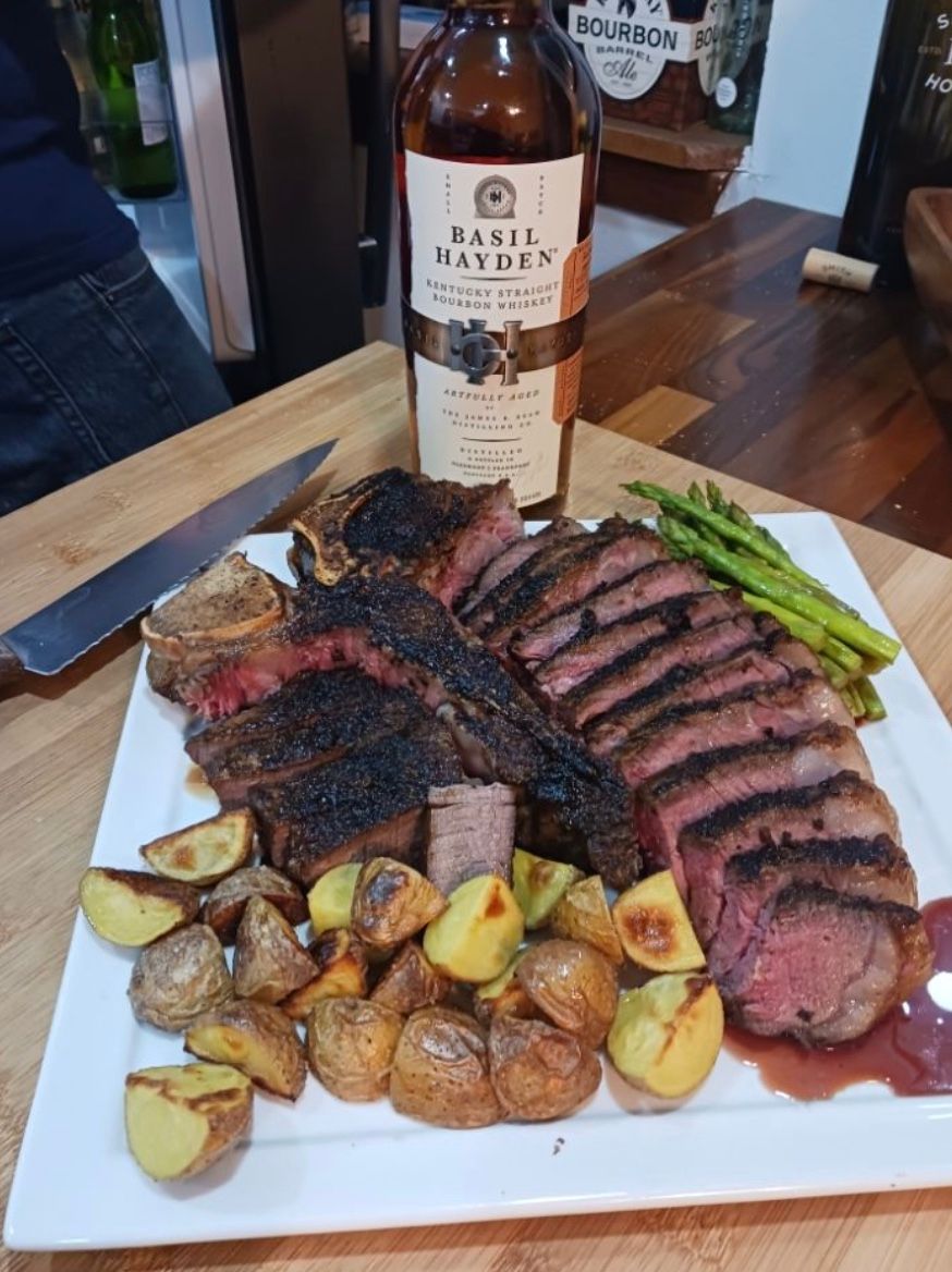 Plated medium-rare bone-in steak, thinly sliced on a white platter with roasted baby potatoes and grilled asparagus, served next to a bottle of Basil Hayden bourbon on a wooden kitchen counter