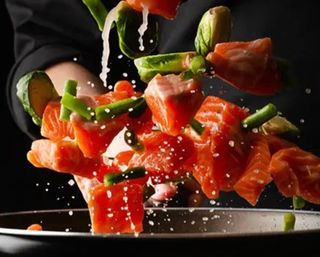 Vibrant cubed salmon and green vegetables (green beans and Brussels sprout halves) mid-air over a frying pan with creamy sauce droplets, dynamic cooking action on a dark background