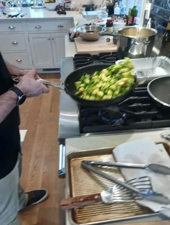 Hands tossing chopped Brussels sprouts in a black skillet over a gas stove in a bright home kitchen, with a baking sheet and tongs on the counter