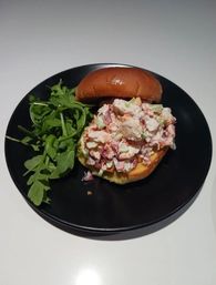 Creamy lobster-salad roll on a glossy brioche bun served with fresh arugula on a black plate over a white tabletop.