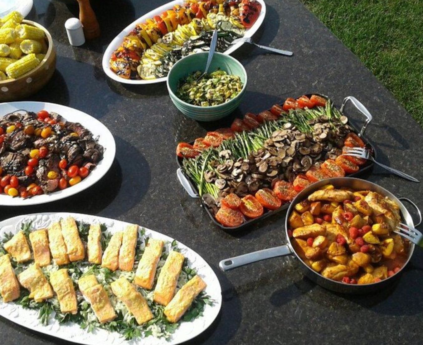 Outdoor backyard summer buffet on a dark stone table with colorful platters of grilled vegetables (asparagus, mushrooms, roasted tomatoes), seared salmon on greens, sliced steak with cherry tomatoes, corn on the cob, a green salad bowl, and a pan of roasted potatoes and chicken.