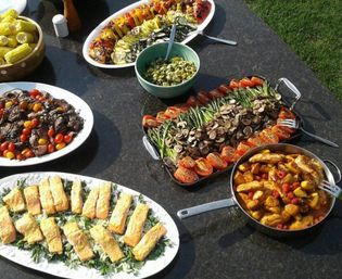 Outdoor backyard summer buffet on a dark stone table with colorful platters of grilled vegetables (asparagus, mushrooms, roasted tomatoes), seared salmon on greens, sliced steak with cherry tomatoes, corn on the cob, a green salad bowl, and a pan of roasted potatoes and chicken.