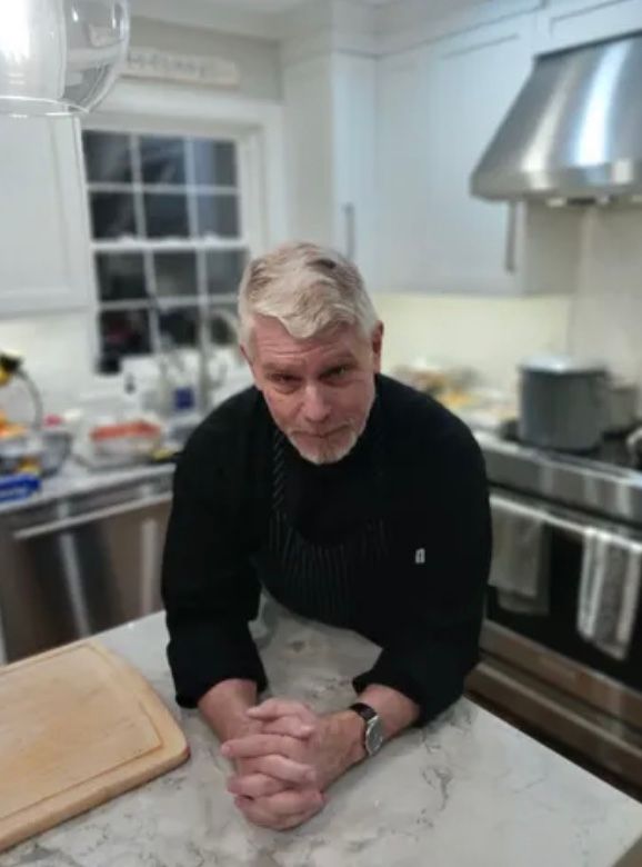 Gray‑haired man in a black chef’s jacket leaning on a marble kitchen island with a cutting board, stainless‑steel range and hood, and a windowed backsplash in a bright modern home kitchen.