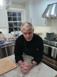 Gray‑haired man in a black chef’s jacket leaning on a marble kitchen island with a cutting board, stainless‑steel range and hood, and a windowed backsplash in a bright modern home kitchen.