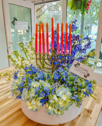 Festive Hanukkah floral centerpiece featuring a brass menorah with nine tall orange-pink tapered candles surrounded by blue and white flowers in a round white vase on a wooden table by a shop window.