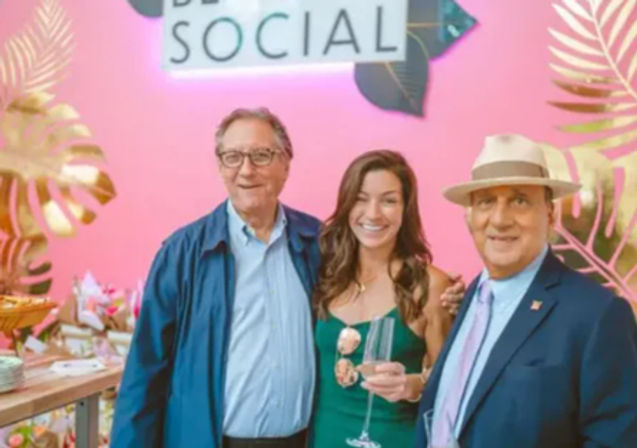 Group photo at a social event with a bright pink backdrop and gold tropical leaf decor; a woman in a green dress holds a champagne flute while two men in a jacket and a suit with a fedora smile beside her.
