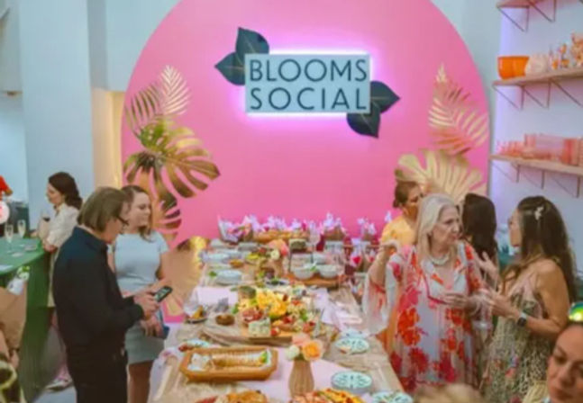 Chic indoor floral-themed social event in a pink arched room with tropical leaf décor, a long table of colorful appetizers and desserts, and guests mingling.