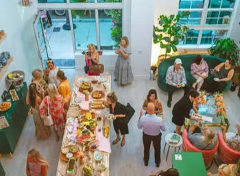 Overhead view of a bright modern urban event space with large windows — cheerful guests mingling around a long buffet table of colorful platters and desserts, green sofa seating and potted plants for a casual daytime gathering.