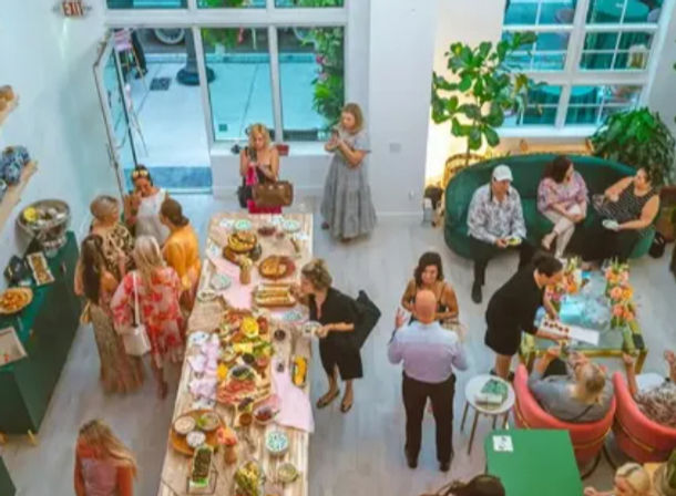 Overhead view of a bright modern urban event space with large windows — cheerful guests mingling around a long buffet table of colorful platters and desserts, green sofa seating and potted plants for a casual daytime gathering.