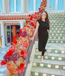 Person in black outfit and pink sunglasses posing on colorful tiled Mediterranean-style outdoor stairs next to a railing wrapped in red and pink floral garland and a terracotta-roofed building with columns