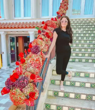 Person in black outfit and pink sunglasses posing on colorful tiled Mediterranean-style outdoor stairs next to a railing wrapped in red and pink floral garland and a terracotta-roofed building with columns