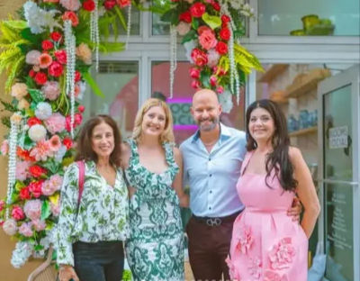 Four people smiling and posing under a vibrant pink-and-red floral arch at a boutique storefront, dressed in colorful summer outfits for a celebratory group photo.