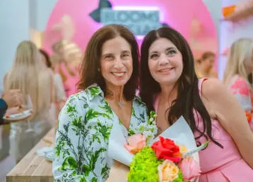 Two smiling women posing at a lively indoor event with a pink backdrop, one holding a vibrant bouquet of red, pink and green flowers.