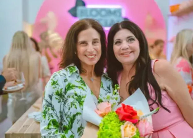 Two smiling women posing at a lively indoor event with a pink backdrop, one holding a vibrant bouquet of red, pink and green flowers.