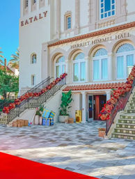 Sunlit Mediterranean-style building entrance with arched windows, twin staircases draped in red floral garlands, potted plants and a patterned tiled plaza.