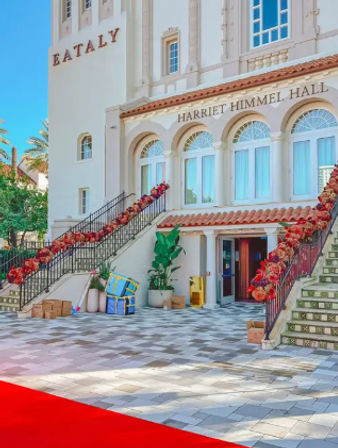 Sunlit Mediterranean-style building entrance with arched windows, twin staircases draped in red floral garlands, potted plants and a patterned tiled plaza.