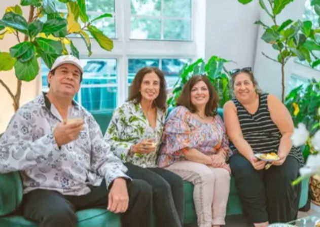 Four adults smiling on a sofa at a casual indoor gathering with drinks and snacks, surrounded by large houseplants and sunlit windows.