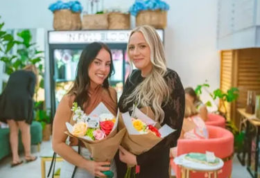 Two smiling women holding vibrant bouquets of roses and mixed blooms inside a bright floral studio with pink lounge chairs and lush green plants