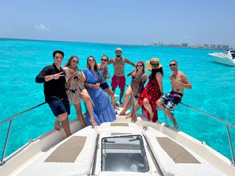 Happy group on the bow of a white yacht enjoying a boat-party vacation over vivid turquoise tropical water with a resort-lined coastline and clear blue sky