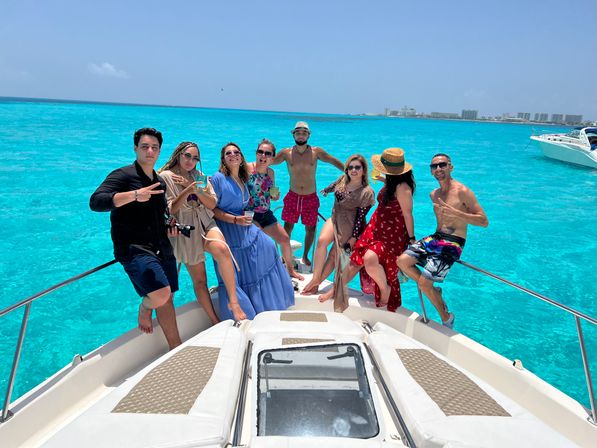 Happy group on the bow of a white yacht enjoying a boat-party vacation over vivid turquoise tropical water with a resort-lined coastline and clear blue sky