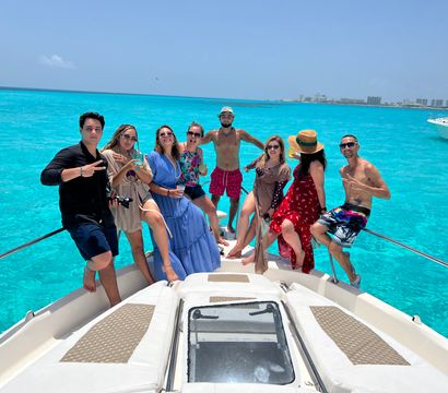 Happy group on the bow of a white yacht enjoying a boat-party vacation over vivid turquoise tropical water with a resort-lined coastline and clear blue sky
