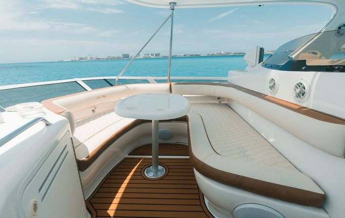 Luxury yacht deck with curved cream-and-tan cushioned seating around a small white table, teak flooring and turquoise ocean with distant coastline under a sunny sky.