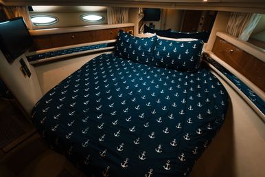 Cozy yacht cabin bedroom with navy anchor-patterned duvet and pillows, oval porthole windows, wood cabinetry and compact TV