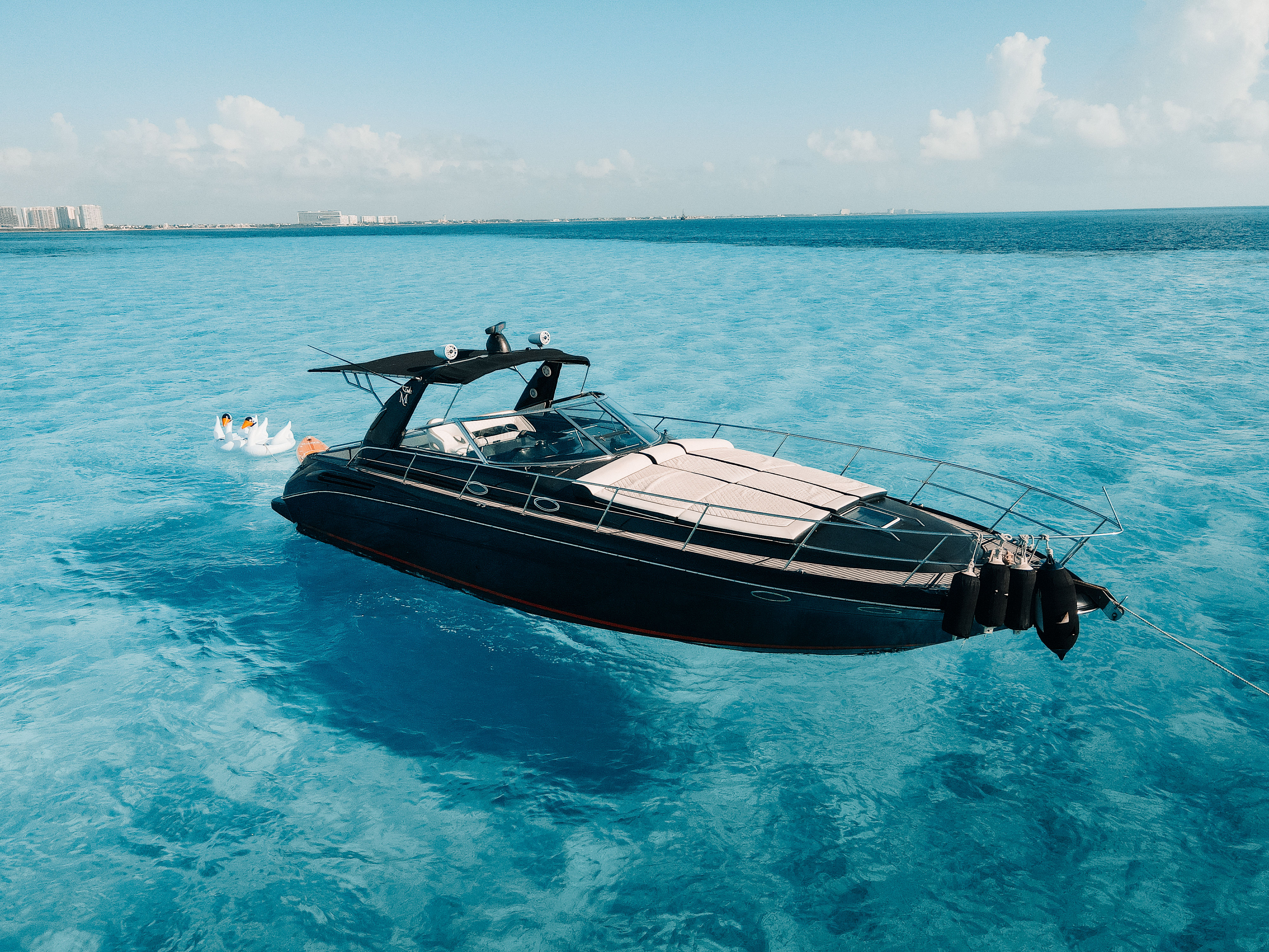 Aerial shot of a sleek black luxury yacht anchored on crystal-clear turquoise water, with inflatable swan floats bobbing nearby and a distant coastal skyline under a sunny blue sky.