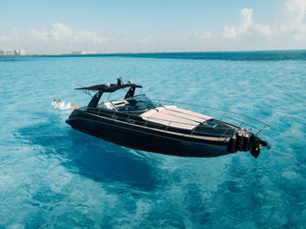 Aerial shot of a sleek black luxury yacht anchored on crystal-clear turquoise water, with inflatable swan floats bobbing nearby and a distant coastal skyline under a sunny blue sky.