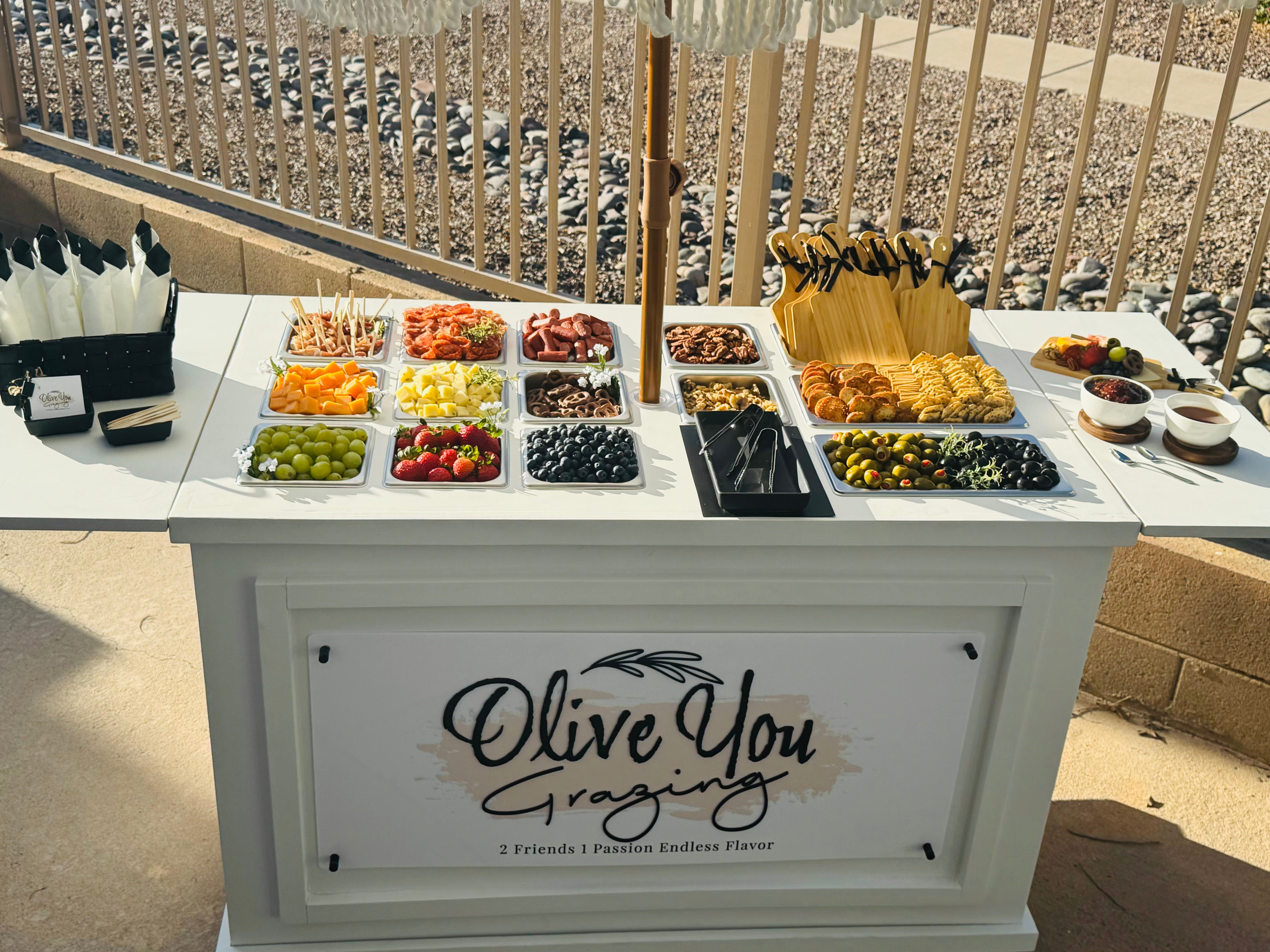 Backyard patio grazing station on a white cart with colorful trays of grapes, strawberries, blueberries, melon, pineapple, olives, crackers, cheeses, charcuterie, wooden boards and serving utensils under a market umbrella