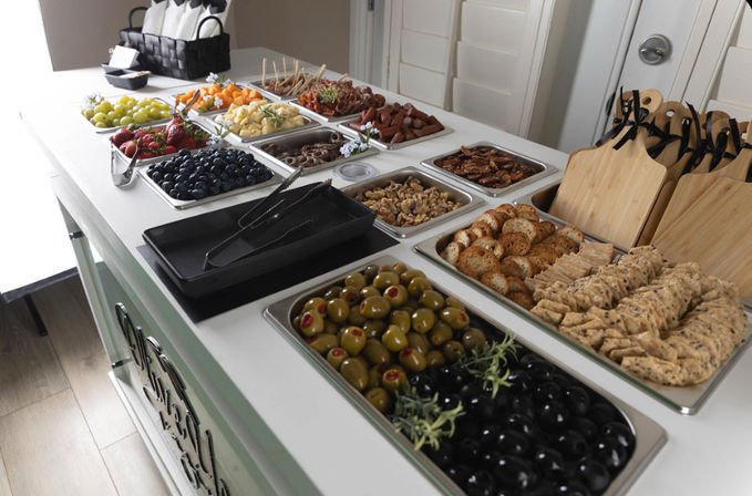 Self-serve charcuterie buffet on white island with trays of green and black olives, assorted crackers and crostini, blueberries, strawberries, grapes, cubed cheeses, nuts, pretzels and cured meats — stylish catered appetizer spread