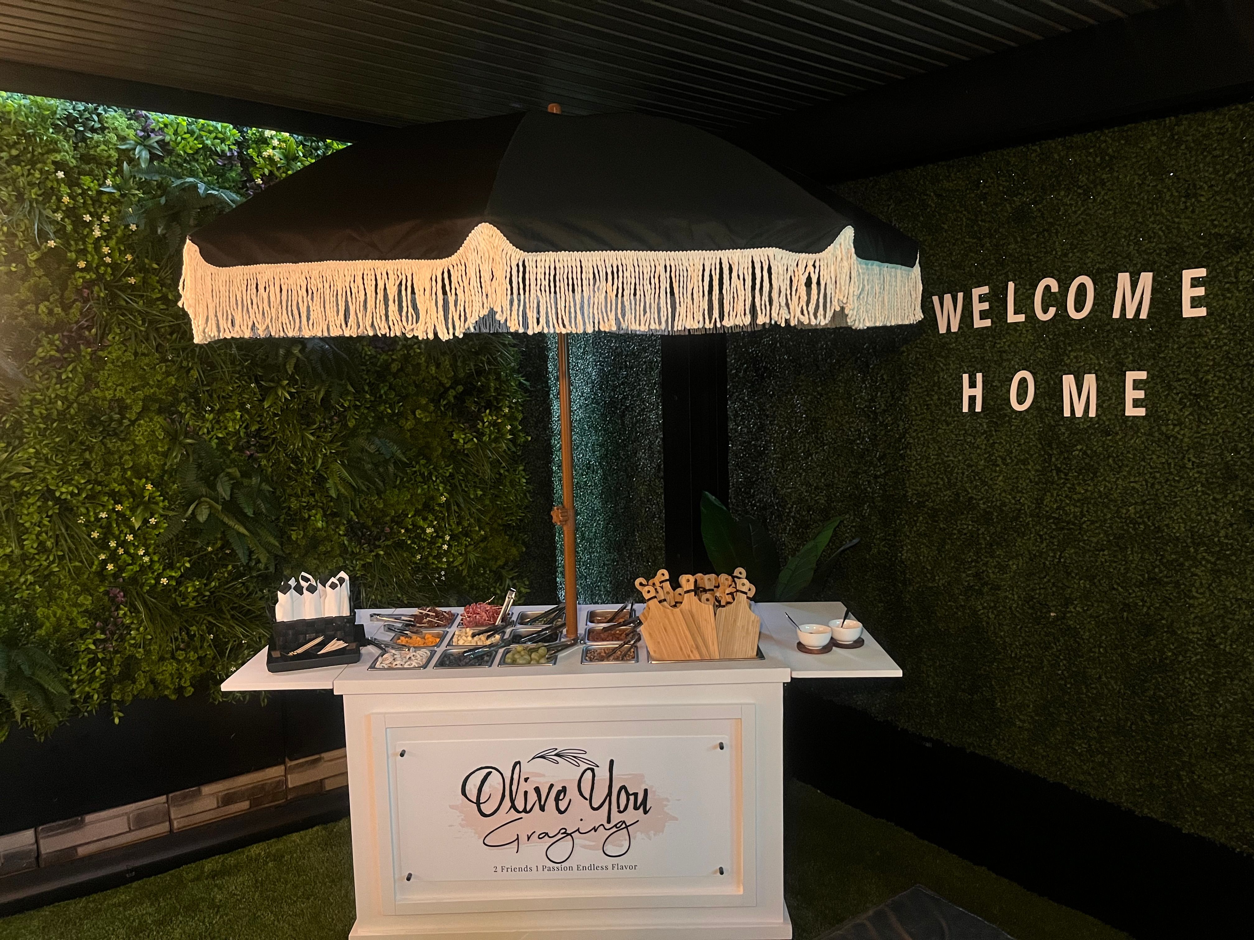 Fringed black umbrella over a white grazing cart loaded with charcuterie trays and serving utensils, set against a lush green wall with “WELCOME HOME” letters in a cozy indoor garden-style space.