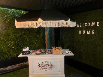 Fringed black umbrella over a white grazing cart loaded with charcuterie trays and serving utensils, set against a lush green wall with “WELCOME HOME” letters in a cozy indoor garden-style space.