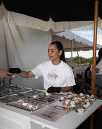 Server at an outdoor food market booth handing a small appetizer cup to a customer, black-gloved hands and rows of fruit and charcuterie tasting cups on the counter under a fringed umbrella.