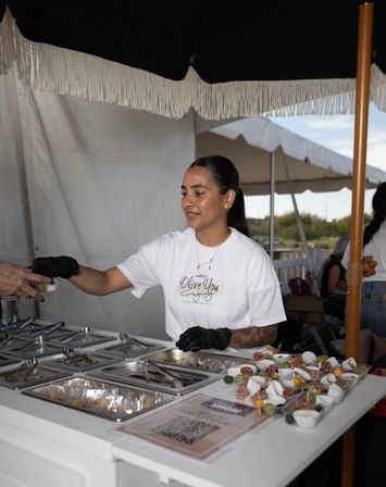 Server at an outdoor food market booth handing a small appetizer cup to a customer, black-gloved hands and rows of fruit and charcuterie tasting cups on the counter under a fringed umbrella.