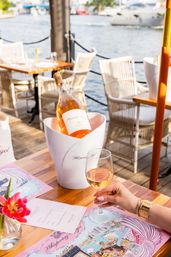 Waterfront dining scene with a chilled bottle of rosé in a white ice bucket on a wooden table, a hand holding a glass of rosé, pink menu and flower, marina and boats in the background.