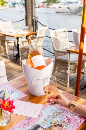 Waterfront dining scene with a chilled bottle of rosé in a white ice bucket on a wooden table, a hand holding a glass of rosé, pink menu and flower, marina and boats in the background.