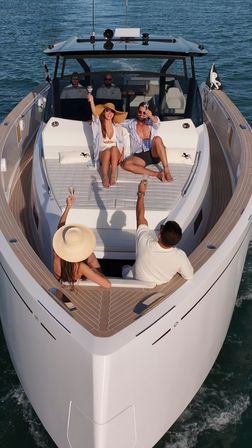Group of friends toasting champagne on the sunlit bow of a luxury yacht with teak decking and crew at the helm in calm coastal waters.