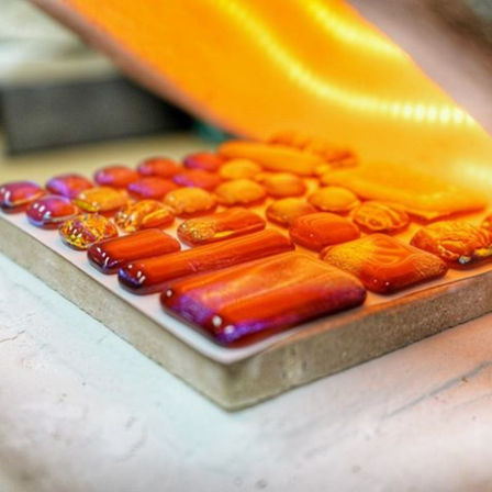 Vibrant orange-red fused glass cabochons and rectangular tiles arranged on a firing board beneath a glowing sheet — artisan mosaic and jewelry components