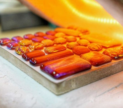 Vibrant orange-red fused glass cabochons and rectangular tiles arranged on a firing board beneath a glowing sheet — artisan mosaic and jewelry components