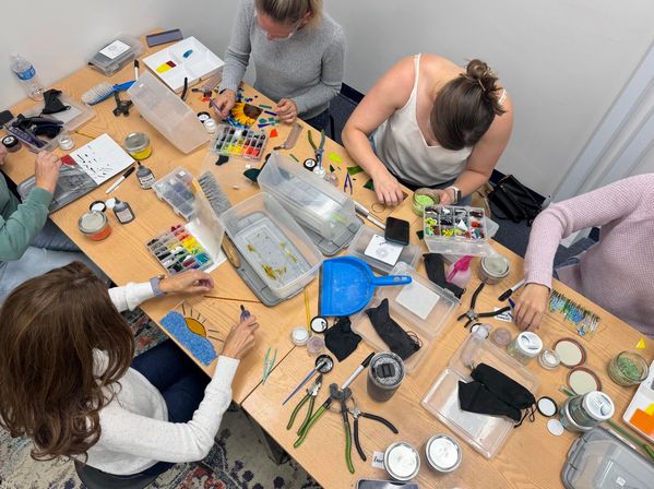 Overhead view of a busy DIY craft workshop: four people working on colorful glass mosaics and jewelry at a wooden table strewn with pliers, tweezers, small containers of glass tiles, jars, and craft tools.