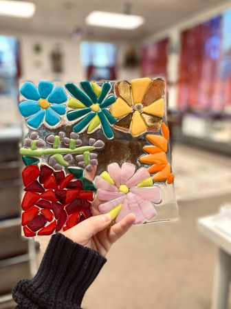 Hand holding a bright fused-glass flower mosaic tile with blue, teal, yellow, orange, red, and pink petals against a blurred art studio background