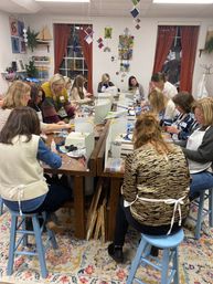Busy art studio workshop with a group of women seated around a long table making mosaic and stained-glass crafts, wearing aprons amid tools, colorful tiles, and hanging glass artwork