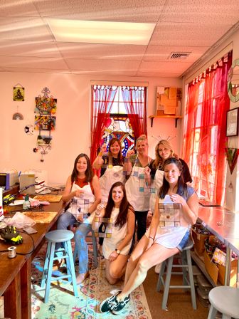 Six women in a sunlit art studio posing with handcrafted stained-glass panels during a DIY stained-glass workshop, with red curtains, worktables, tools and colorful wall art in the background.