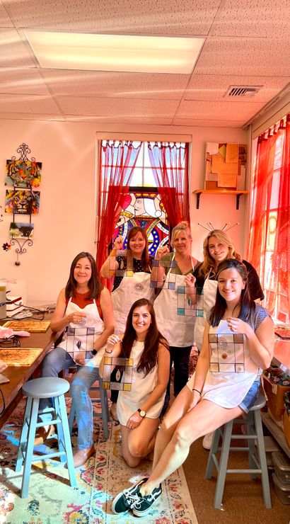 Six women in a sunlit art studio posing with handcrafted stained-glass panels during a DIY stained-glass workshop, with red curtains, worktables, tools and colorful wall art in the background.