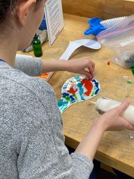 Person assembling a colorful glass-tile mosaic on a spoon-shaped base at a wooden workbench, using a squeeze bottle of adhesive; orange center with blue, green and mirrored tiles.