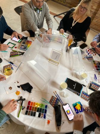 Top-down view of an indoor glass mosaic workshop: participants around a round table arranging colorful tiles, tools, smartphones and drinks in a hands-on craft session.
