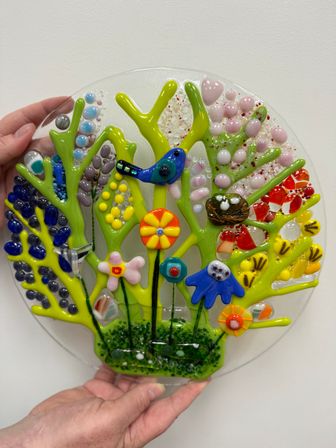 Handheld round fused glass wall art with lime-green tree branches, vibrant textured flowers, a blue bird and nest, colorful beads and dots on a clear background — playful decorative glass panel.